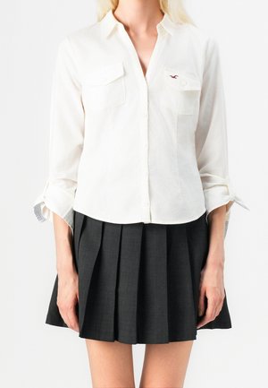 Femme portant une chemise blanche à manches longues boutonnée avec des poches poitrine et une mini-jupe plissée noire, debout les bras détendus.