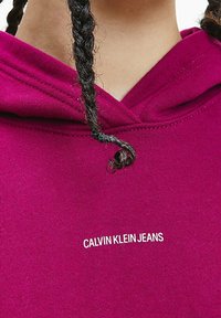Magenta hoodie med dragsko, med en vit "CALVIN KLEIN JEANS" logo. Tillverkad av mjukt material med en slät yta.