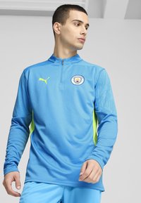 Lichtblauwe sporttop met lange mouwen en hoge kraag, voorzien van een levendig geel logo en zijkanten. Gevoelig materiaal met een grafisch patroon op de mouwen.