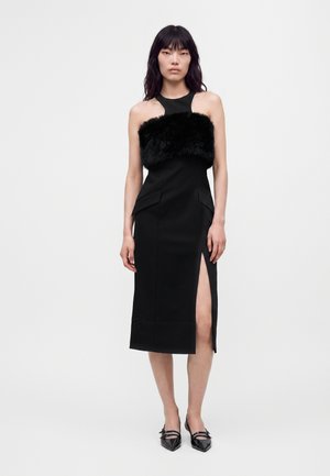 Femme portant une robe midi noire sans manches avec un panneau en fourrure sur la poitrine et une fente haute à la cuisse, associée à des chaussures pointues noires.
