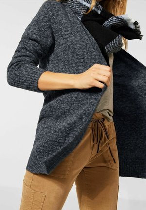 Cardigan tricoté gris charbon avec des manches côtelées, à ouverture frontale et à coupe décontractée. Associé à un pantalon beige avec cordon de serrage adjustable.