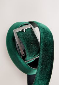 Cintura in velluto verde con fibbia metallica argento, caratterizzata da una finitura strutturata e bordi lisci, arrotolata per l'esposizione.