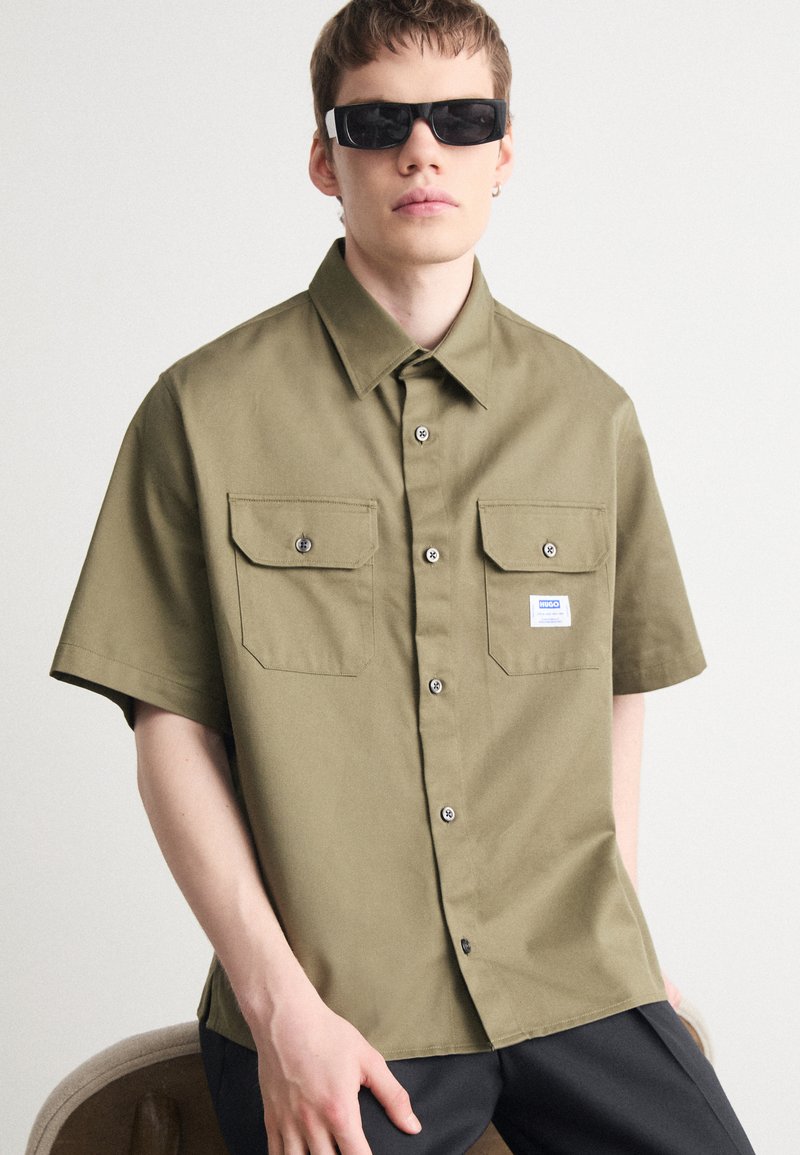 Olive green short-sleeve shirt with two front pockets, buttoned closure, and brand label. Model wears black sunglasses. Smooth fabric texture.