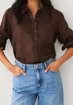 Femme portant une chemise marron à manches longues boutonnée et un jean taille haute bleu clair, les mains dans les poches, debout contre un mur uni.