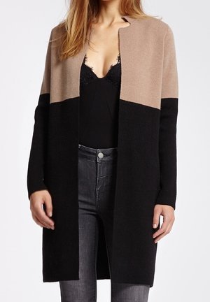 Mujer que lleva un cárdigan largo bicolor en beige y negro sobre una camiseta negra con encaje y jeans gris oscuro, de pie frente a un fondo liso.