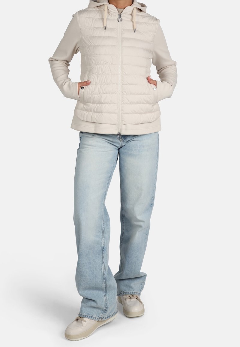 Chaleco acolchado beige con capucha, cierre delantero y bolsillos laterales, combinado con jeans de pierna recta azul claros y zapatillas beige.