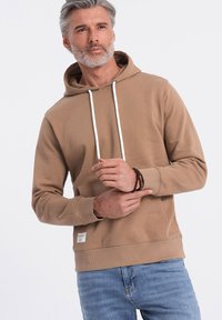 Felpa color sabbia con tasca frontale, cordini bianchi, polsini a coste e un piccolo etichetta sul bordo. Abbinata a jeans blu.