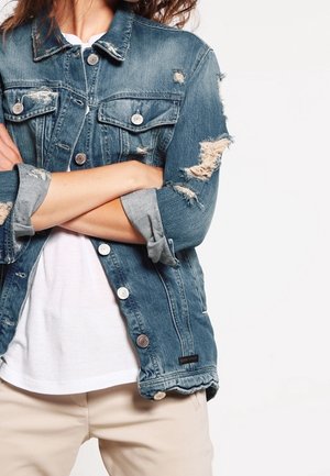 Persoon met gekruiste armen die een versleten blauwe spijkerjack draagt over een wit shirt en beige broek tegen een effen achtergrond.