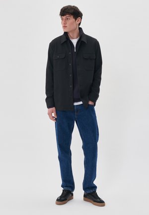 Cămașă neagră cu buton și două buzunare pe piept, purtată deasupra unei bluze navy cu fermoar, combinată cu blugi și adidași negri. Fundal simplu.