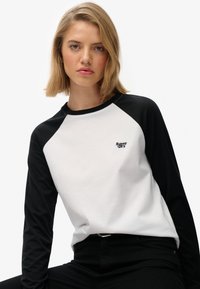 Koszula z długim rękawem typu raglan z białą częścią główną i czarnymi rękawami; posiada małe czarne logo "Superdry" na klatce piersiowej. Wykonana z miękkiego materiału bawełnianego.