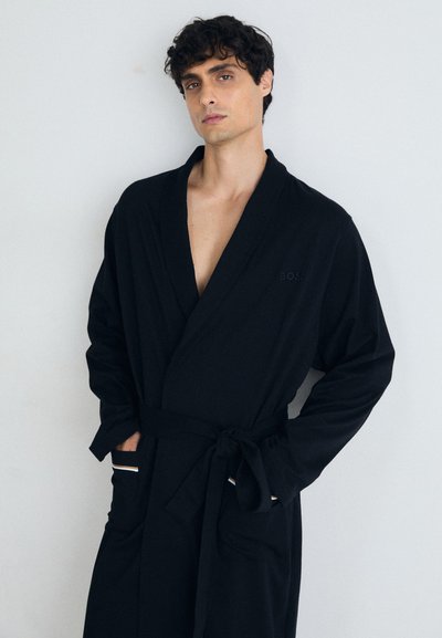 Peignoir noir avec un col châle, ceinture à nouer et poches latérales. Présente un logo discret et une texture lisse et douce.