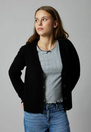 Jeune femme en cardigan noir boutonné sur un haut gris et un jean bleu, regardant sur sa gauche contre un fond gris uni.