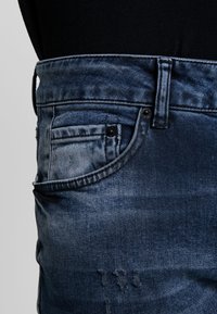 Nära bild av en ficka på blå denimbyxor med sömnadsdetaljer och lätt slitna partier, bärs med en svart topp.