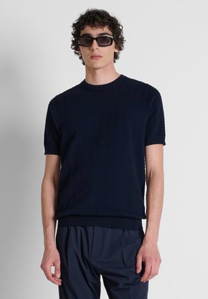 Giovane uomo con capelli ricci che indossa occhiali da sole scuri, una camicia blu navy a maniche corte con trama e pantaloni blu navy su uno sfondo uniforme.