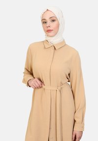 Vestido beige de botonadura con cuello, mangas largas y un cinturón a juego en la cintura, hecho de una tela ligera y texturizada.