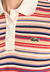 Pruhovaná polo košile v červené, modré a krémové barvě, s jemnou strukturou. Má bílý límec a tři knoflíky na léze. Vyšívané logo Lacoste.