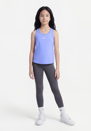 Fille debout en débardeur Nike violet, leggings gris foncé, chaussettes Nike blanches et baskets blanches sur un fond blanc uni.