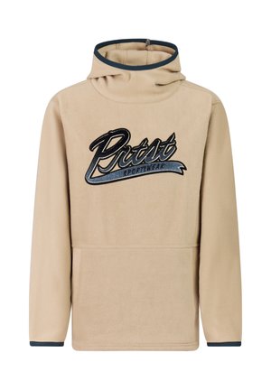 Jongen met beige hoodie met de tekst "Rist", donkerblauwe broek, zwarte schoenen en een blauw gestreepte muts, staand tegen een effen achtergrond.