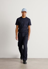 T-shirt nera a maniche corte con texture e pantaloni neri coordinati, abbinati a un cappellino blu chiaro e calzature nere, su uno sfondo neutro.