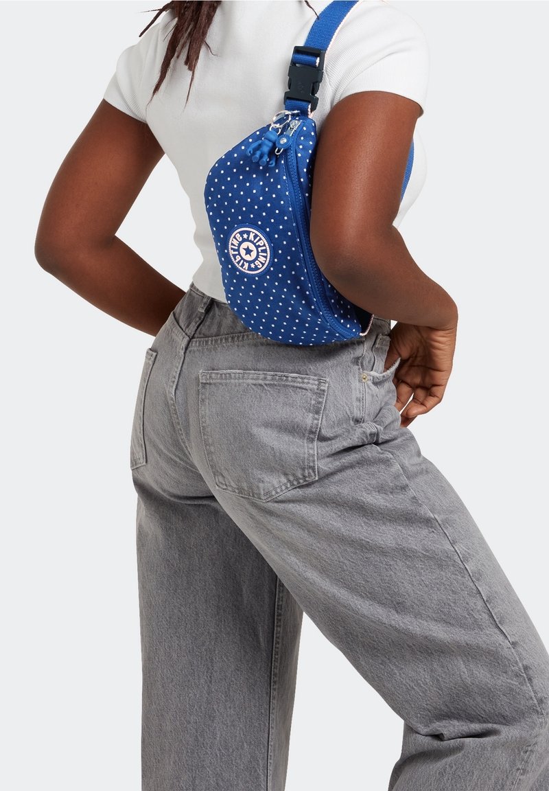 Femme portant un haut blanc et un jean gris porte un sac bandoulière bleu à pois sur son dos, la main droite sur la hanche.