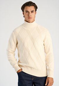 Roomkleurige coltrui met kabelgebreid patroon, met textuurrijke diamantmotieven en geribbelde manchetten, gecombineerd met donkere jeans.