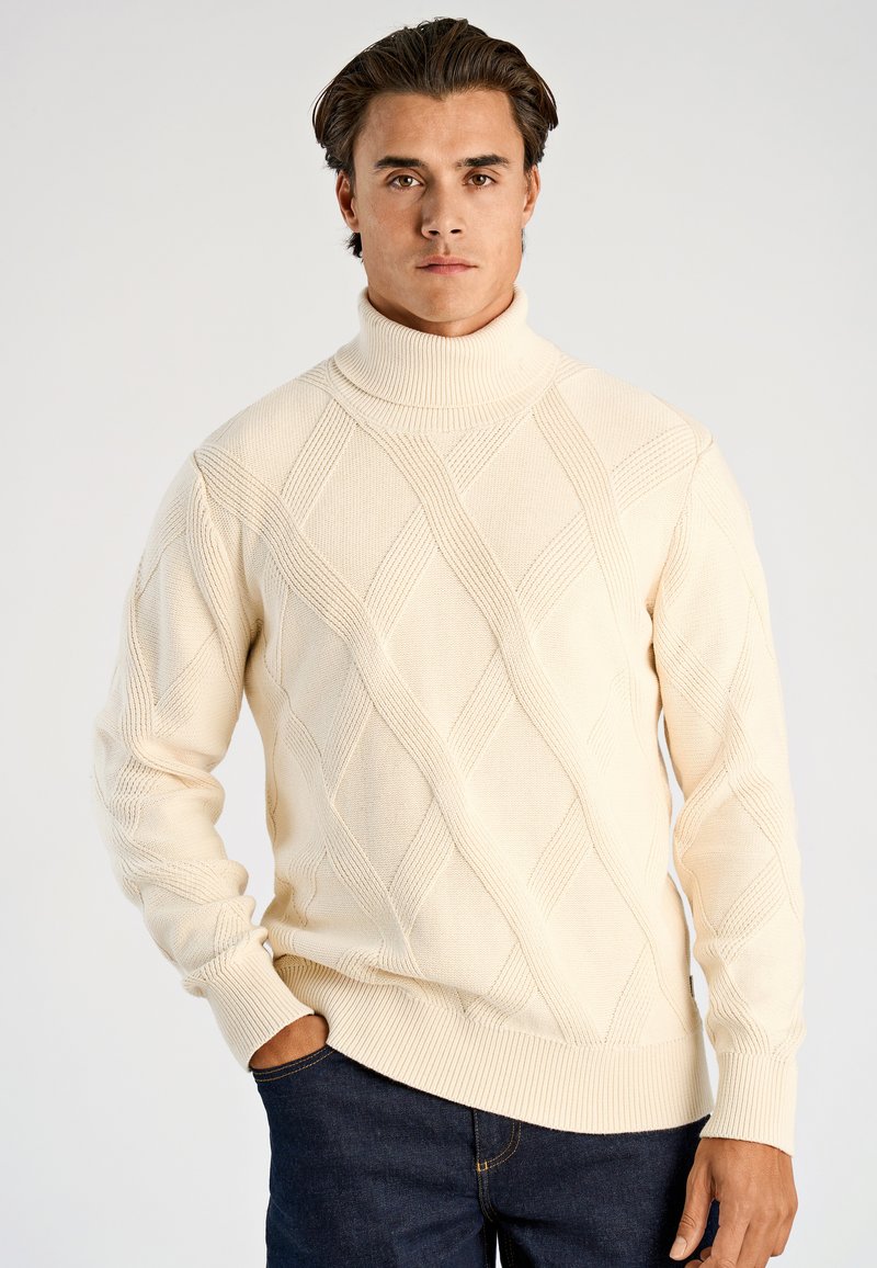 Roomkleurige coltrui met kabelgebreid patroon, met textuurrijke diamantmotieven en geribbelde manchetten, gecombineerd met donkere jeans.