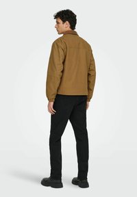 Moški s kodrastimi temnimi lasmi, oblečen v rjav plašč, črne hlače in črne čevlje, stoji obrnjeno stran od kamer na enobarvni svetli podlagi.