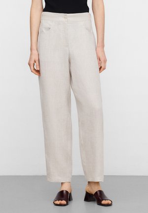 Frau trägt hochtaillierte, hellbeige Hosen mit weitem Bein und Fronttaschen sowie dunkelbraune offene, hohe Sandalen und steht auf grauem Boden.