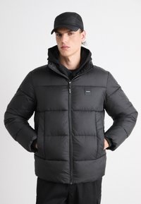 Svart boblejakke med glidelås foran, med quiltet design, høy krage og to sidelommer; brukt over en svart hettegenser.