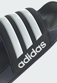 Black slide sandal featuring white three-stripe design and logo. Materials include rubberized strap and textured footbed surface.