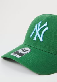 Grønn baseballcap med en blå brodert "NY"-logo foran, med en buet skjerm og en svart etikett på siden.
