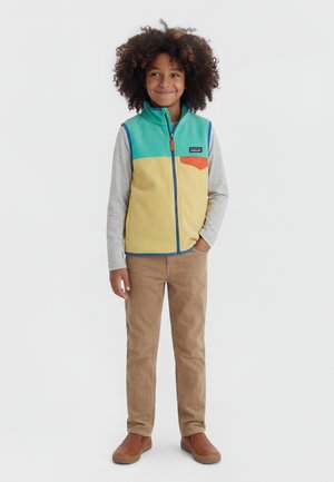 Bambino con capelli ricci che indossa un gilet con zip turchese e giallo sopra una maglia grigia a maniche lunghe, pantaloni marroni e scarpe marroni senza lacci, in piedi con le mani nelle tasche.