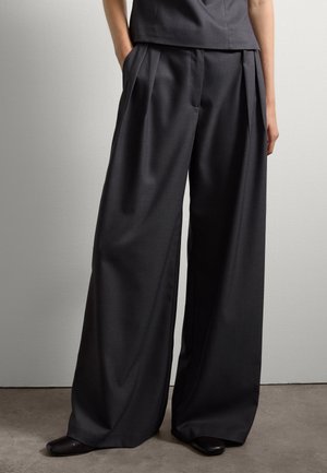 Persoon in hoge taille, wijde, donkergrijze broek met plooien, handen in de zakken en zwarte schoenen, staand op een grijze vloer tegen een witte muur.