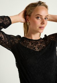 Top de encaje negro con un patrón ondulado, tejido texturizado y bordes festoneados. Los ojos están adornados con pendientes de declaración dorados.