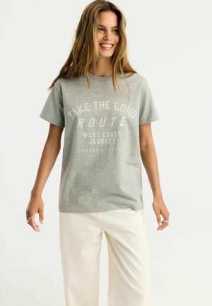 Młoda kobieta z długimi włosami uśmiecha się, nosi szary t-shirt z napisem i białe spodnie na jednolitym jasnym tle.