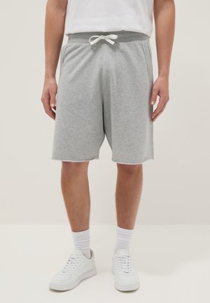 Uomo con pantaloncini grigio chiaro con coulisse, calzini bianchi e scarpe da ginnastica bianche, in piedi su uno sfondo uniforme.