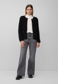 Schwarze flauschige Strickjacke mit goldenen Knöpfen, getragen über einem weißen Rollkragenpullover und kombiniert mit grauen weiten Jeans und schwarzen Plateauschuhen.