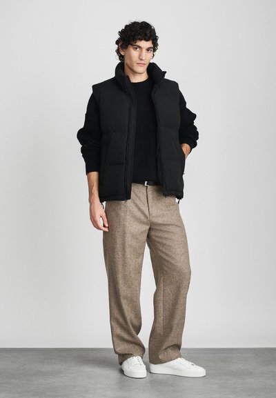 Zwarte gewatteerde vest over een gestructureerd zwart sweater, gecombineerd met lichtbruine op maat gemaakte broek en witte sneakers. Eenvoudig, modern ontwerp.