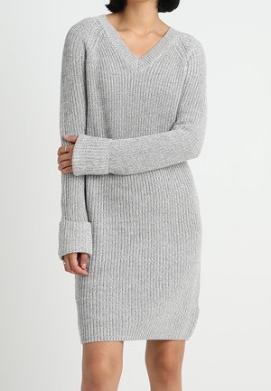 Kobieta w jasnoszarej dzianinowej sukience z długim rękawem, dekoltem w serek i mankietami, stojąca na tle jednolitego białego tła.