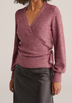 Femme portant un pull cache-cœur mauve avec des paillettes et des manches longues, associé à une jupe en cuir noire taille haute.