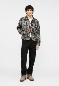 Μπουφάν από τζιν σε μαύρο και γκρι με κουμπιά κλεισίματος, φορεμένο πάνω από ένα λευκό T-shirt, σετ με μαύρο παντελόνι και καφέ μπότες.