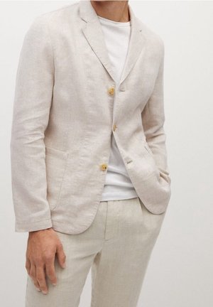 Mann trägt ein beiges Leinen-Sakko mit zwei Knöpfen über einem weißen T-Shirt und passenden beigen Leinenhosen, eine Hand in der Tasche.