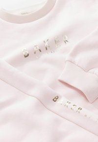 Ljust rosa sweatshirt med ribbad halsringning och fållar. Har metallisk guldig text "BAKER by TED BAKER" som är tydligt utställd. Mjuk tygtextur.
