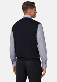 Pánský námořnický bezrukávový svetr vyrobený ze pleteného materiálu, nošený přes světle modrou pruhovanou košili s manžetami na knoflíky, doplněný o tmavé kalhoty.