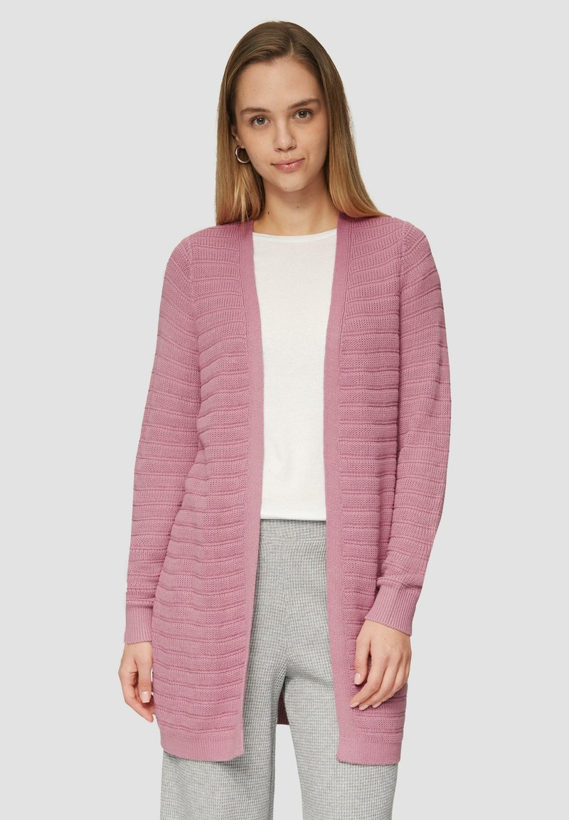 Lichtroze cardigan met een gestructureerd ribpatroon, open voorkant, lange mouwen en zachte gebreide stof. Gemaakt over een witte top en grijze broek.