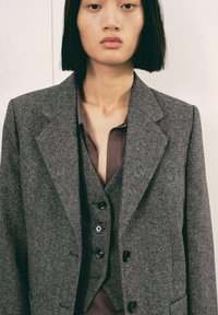 Blazer de lana gris con solapas afiladas, que cuenta con un cierre de dos botones y una tela texturizada. Llevado sobre una camisa marrón oscuro con escote en V.