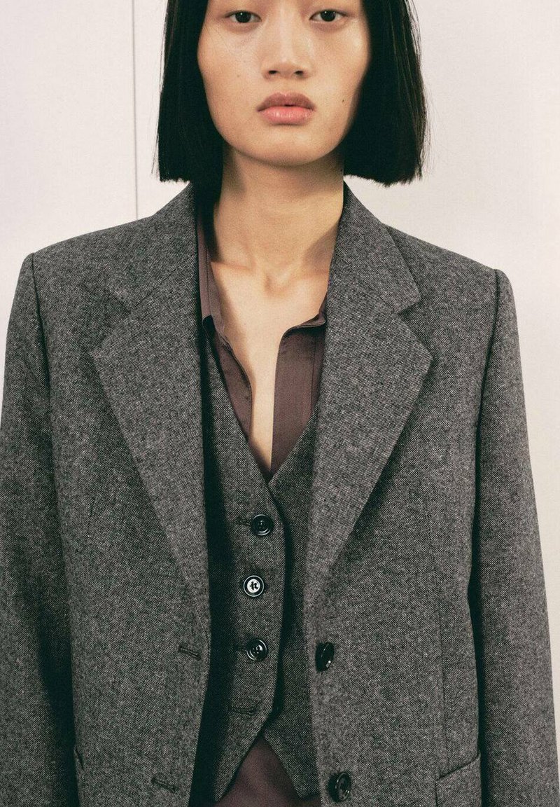 Blazer de lana gris con solapas afiladas, que cuenta con un cierre de dos botones y una tela texturizada. Llevado sobre una camisa marrón oscuro con escote en V.