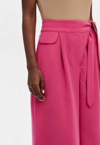Roze op maat gemaakte broeken met een strikceintuur, voorzien van zijzakken en plooidetails, gemaakt van een gladde stof met een matte afwerking.