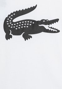 T-shirt blanche avec un grand graphisme de crocodile noir présentant un corps texturé et à motifs, ainsi qu'une queue pointue, positionné sur le côté gauche.