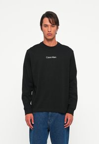 Mand iført en sort langærmet Calvin Klein skjorte og blå jeans, stående mod en ensfarvet hvid baggrund.